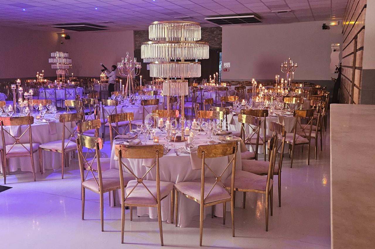 Salle de réception décorée avec des tables rondes, chaises dorées, chandeliers et éclairage tamisé.