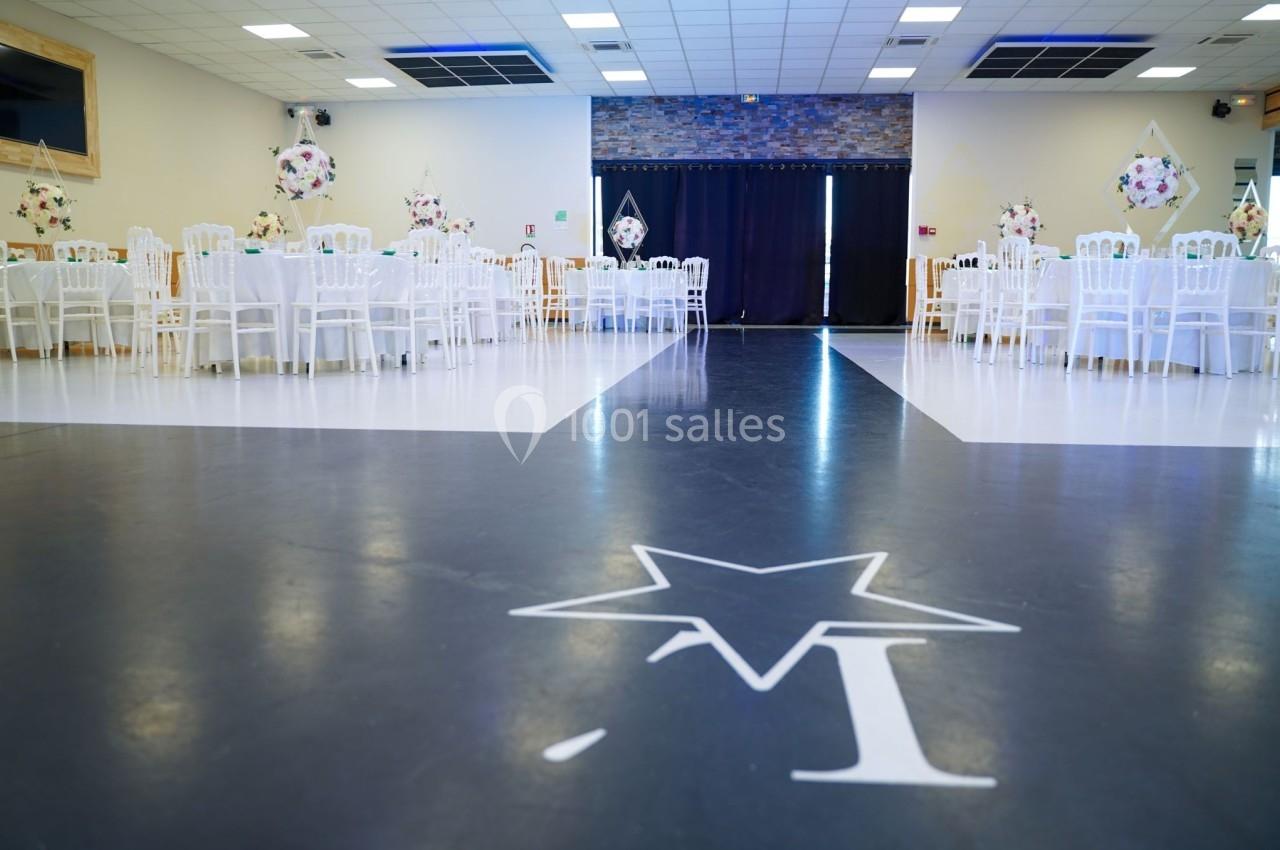Salle de réception décorée avec des tables rondes, chaises blanches et ballons, avec une piste de danse centrale.
