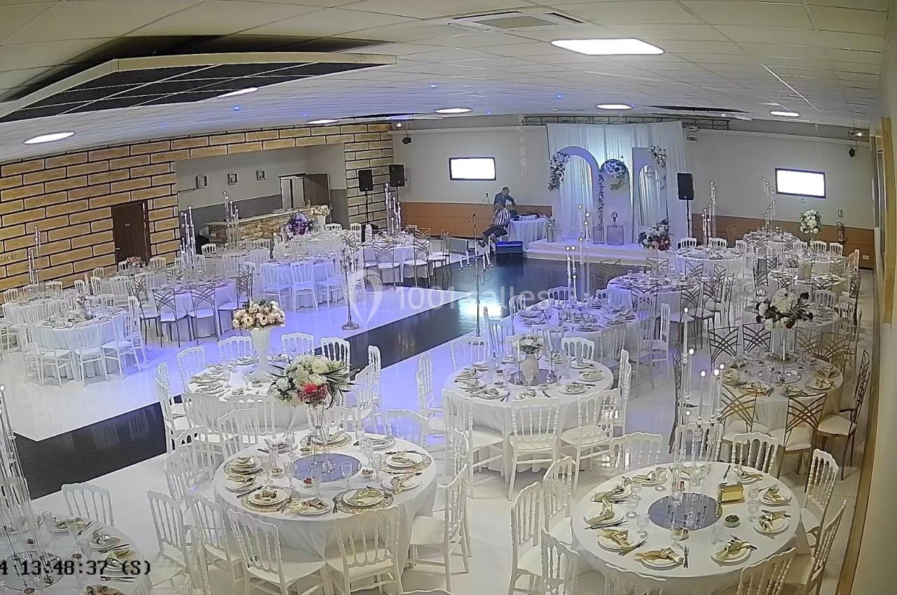 Salle de réception décorée avec des tables rondes dressées, des chaises blanches et une arche fleurie au fond.