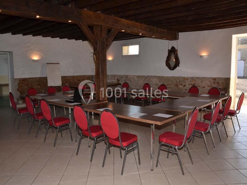 Salle de réunion avec tables disposées en U, chaises rouges, mur en pierre et poutres apparentes.