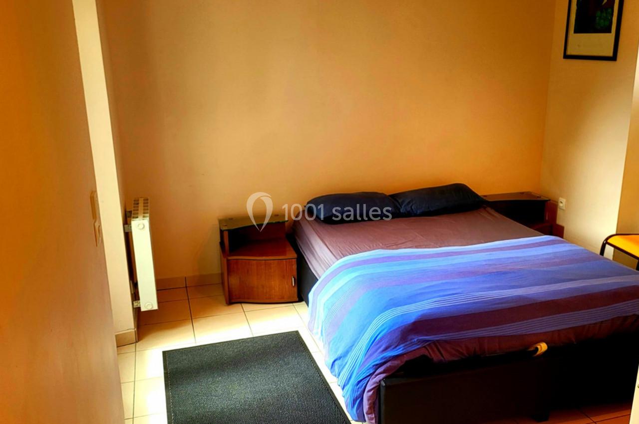 Lit double avec draps bleus dans une chambre simple aux murs clairs, équipée de tables de chevet et d'un radiateur.