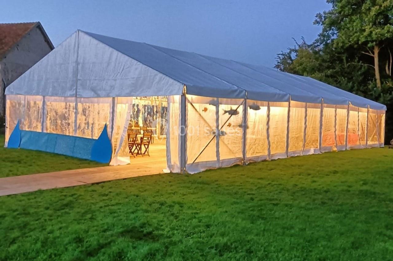 Grande tente éclairée en soirée, installée sur une pelouse, avec des arbres et un bâtiment en arrière-plan.