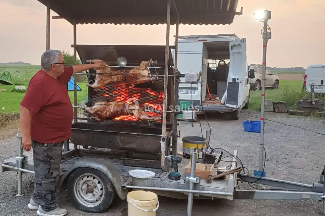 Un homme tourne des broches avec des viandes rôtissant sur un barbecue mobile en plein air, près de véhicules.