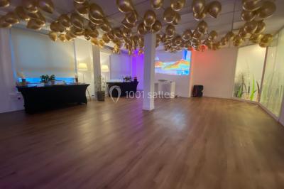 Salle lumineuse avec parquet, plafond décoré de ballons dorés, table avec plantes et vue sur des bâtiments urbains.