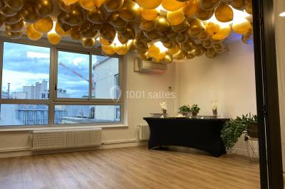 Salle lumineuse avec parquet, plafond décoré de ballons dorés, table avec plantes et vue sur des bâtiments urbains.