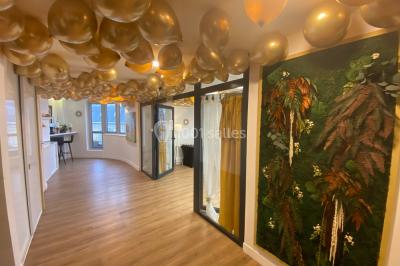 Salle lumineuse avec parquet, plafond décoré de ballons dorés, table avec plantes et vue sur des bâtiments urbains.