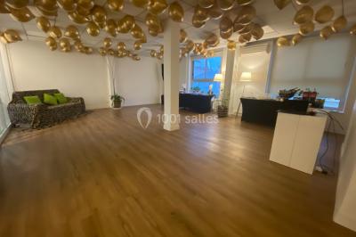 Salle lumineuse avec parquet, plafond décoré de ballons dorés, table avec plantes et vue sur des bâtiments urbains.