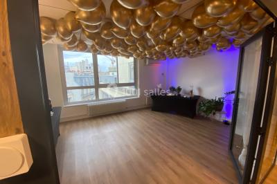 Salle lumineuse avec parquet, plafond décoré de ballons dorés, table avec plantes et vue sur des bâtiments urbains.