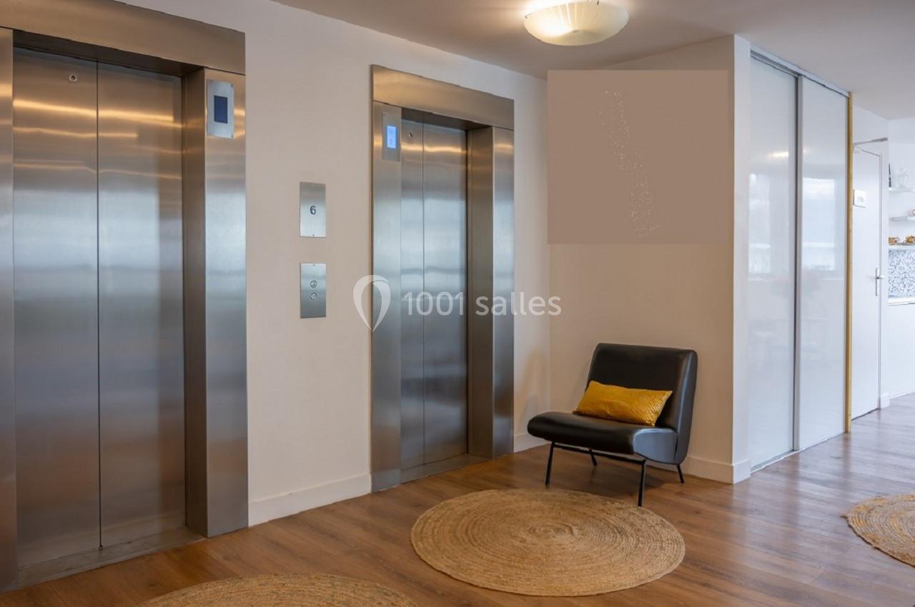 Deux ascenseurs modernes dans un hall lumineux avec un fauteuil noir, un coussin jaune et des tapis ronds au sol.