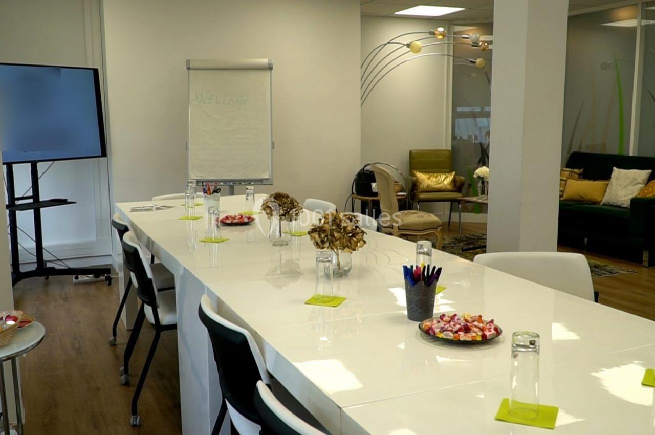 Salle de réunion lumineuse avec une grande table blanche, des chaises, un tableau de conférence et un coin salon en arrière…