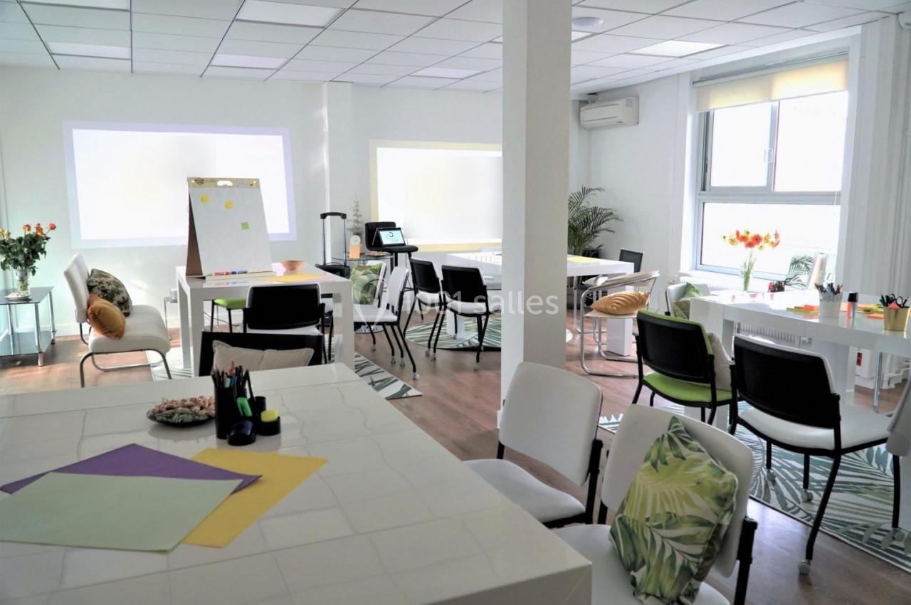 Salle lumineuse aménagée avec des tables, chaises, plantes, un tableau de présentation et des fournitures de bureau.
