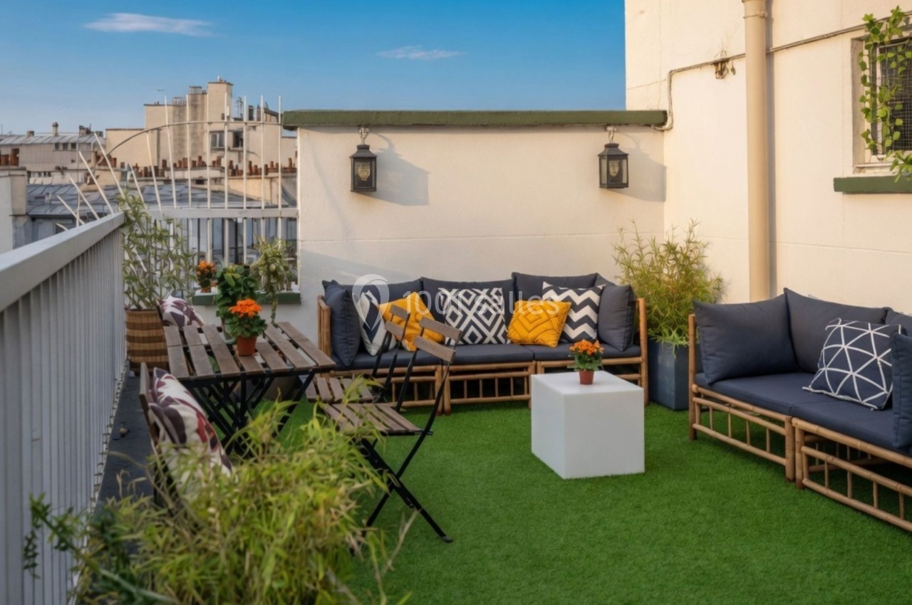Terrasse aménagée avec mobilier en bois, coussins colorés, table, chaises et plantes sur un sol en gazon synthétique.