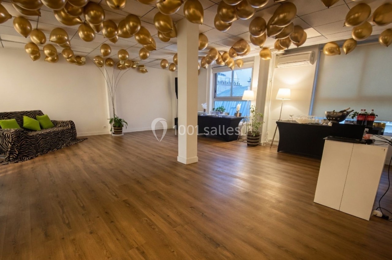 Salle lumineuse avec parquet, canapé à motifs, ballons dorés suspendus et tables pour buffet en arrière-plan.