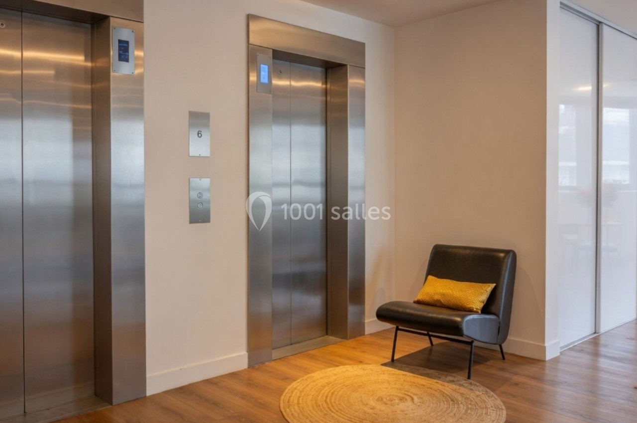 Deux ascenseurs modernes dans un hall lumineux avec un fauteuil noir et un coussin jaune à côté.
