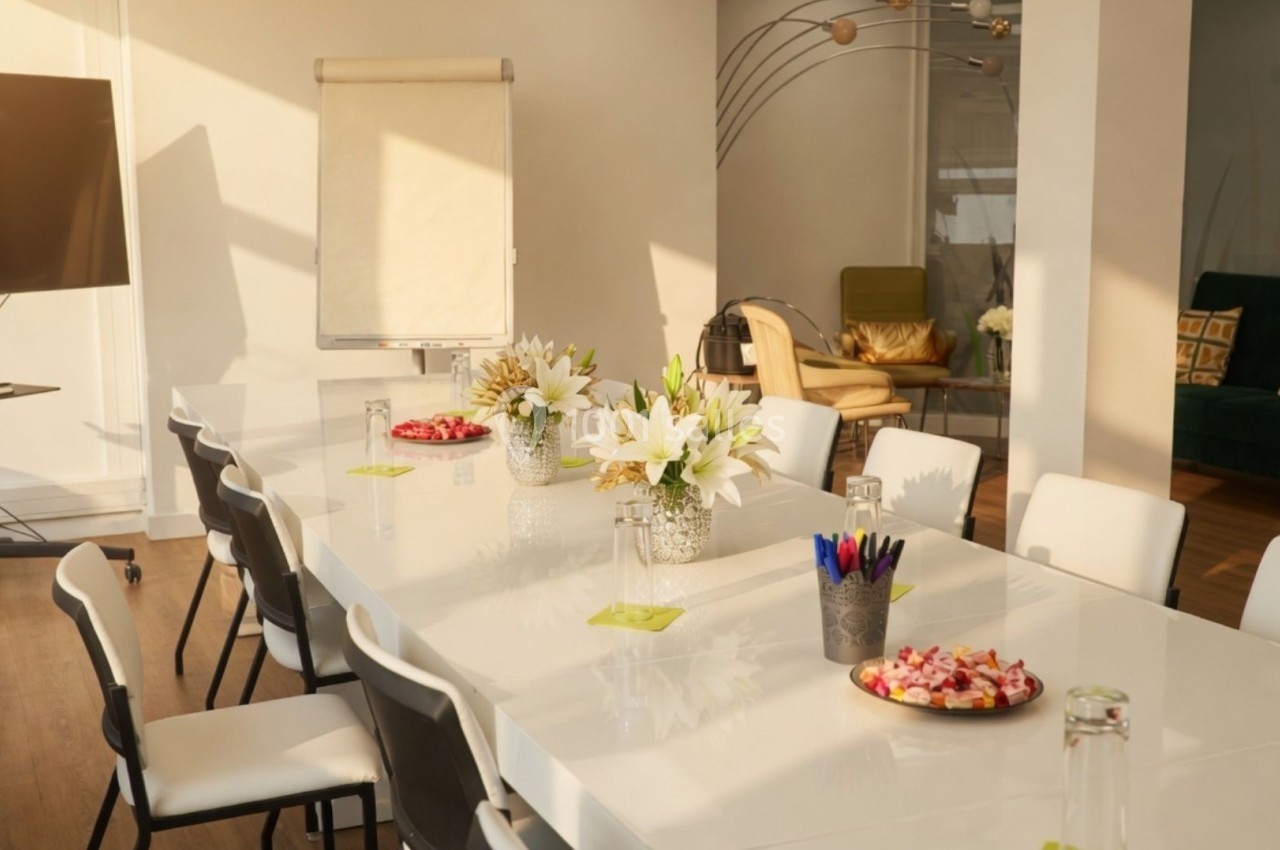 Salle de réunion lumineuse avec une grande table blanche, des chaises, un tableau de conférence et des décorations florales.