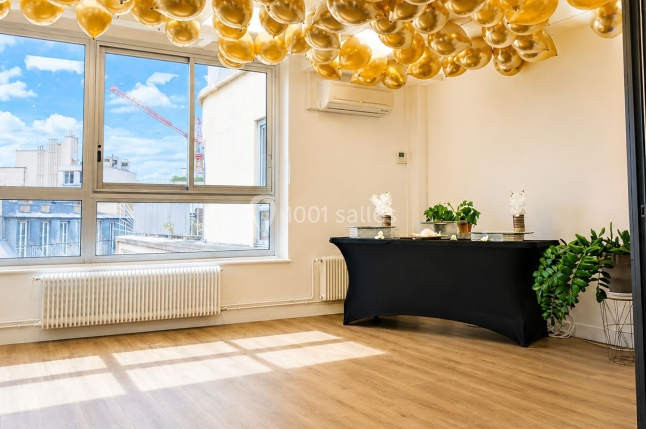 Salle lumineuse avec parquet, décorée de ballons dorés au plafond, une table noire et des plantes près d'une fenêtre.