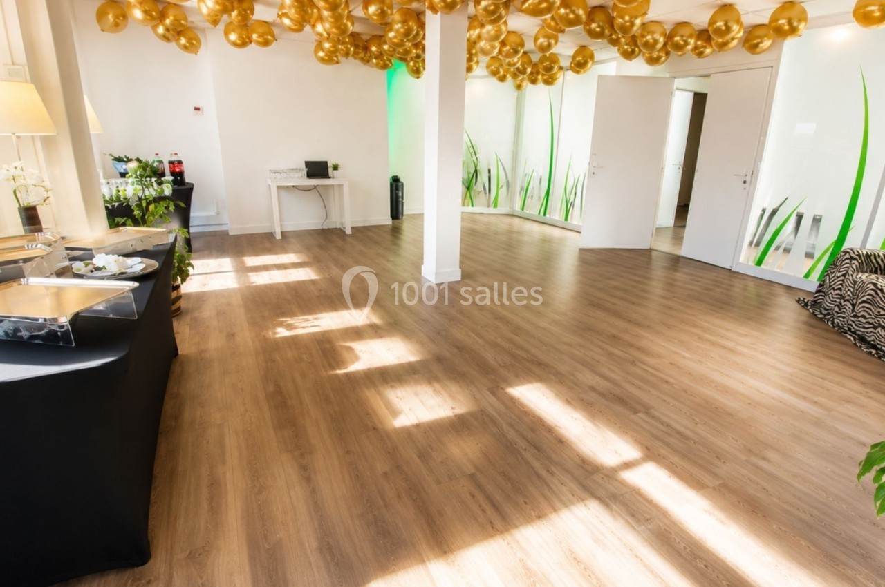 Salle lumineuse avec parquet, décorée de ballons dorés au plafond, tables de buffet et plantes en arrière-plan.