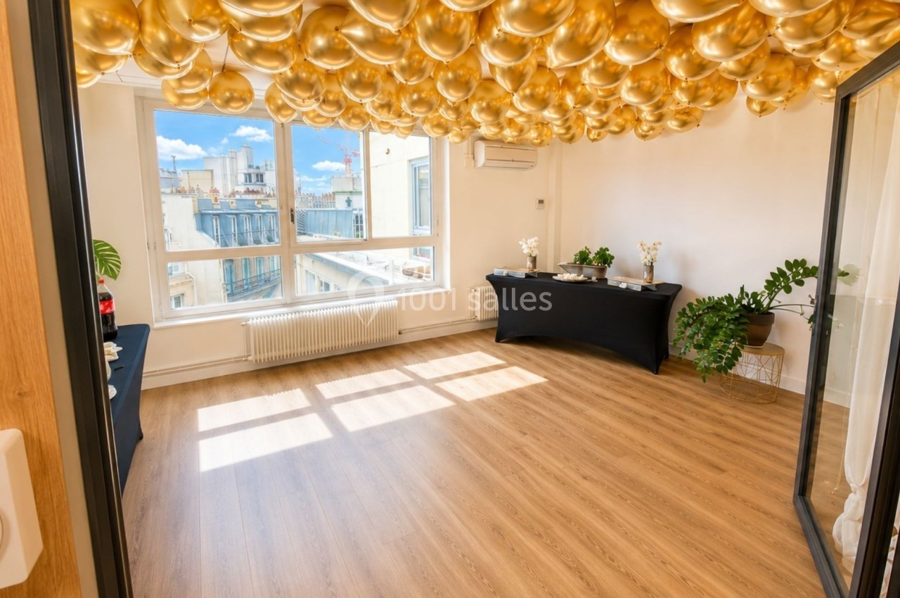 Salle lumineuse avec parquet, décorée de ballons dorés au plafond, tables noires et plantes près des fenêtres.