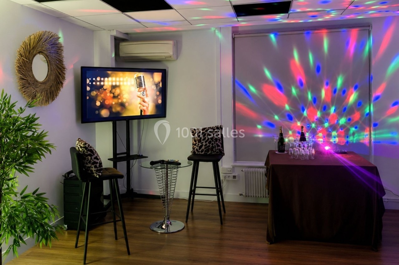 Salle décorée avec des lumières colorées, un écran affichant un micro, des tabourets et une table avec des verres.
