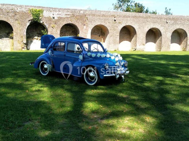 Voiture ancienne bleue décorée de fleurs blanches, garée sur une pelouse devant des arches en pierre.