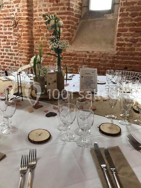 Table décorée pour un événement avec nappes blanches, rondins de bois, fleurs et vaisselle dans une salle en briques.