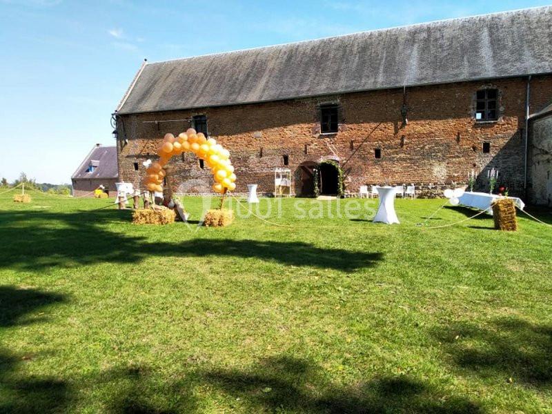 Grande pelouse avec arche de ballons orange, tables hautes et bottes de foin devant un bâtiment en briques anciennes.