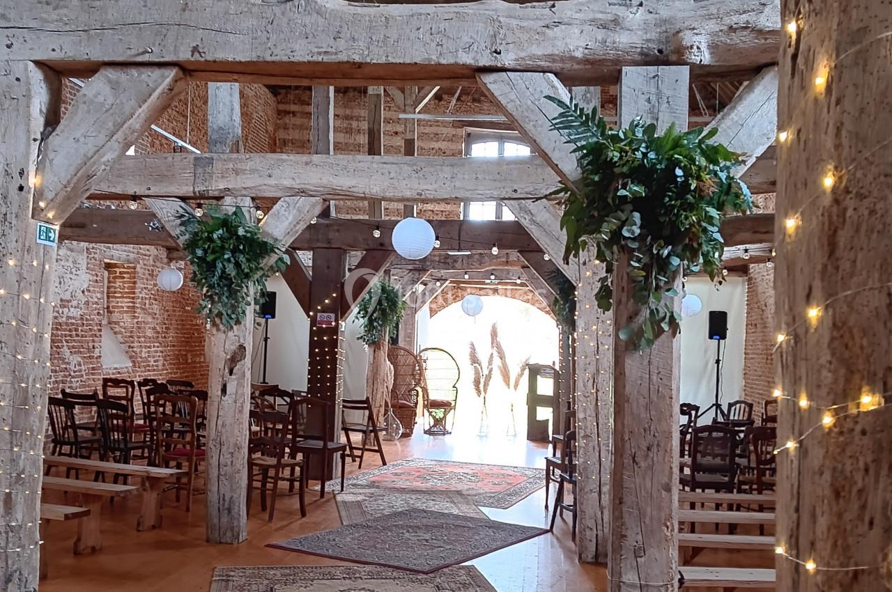 Intérieur d'une grange aménagée avec des poutres en bois, tapis, guirlandes lumineuses et chaises disposées pour un…