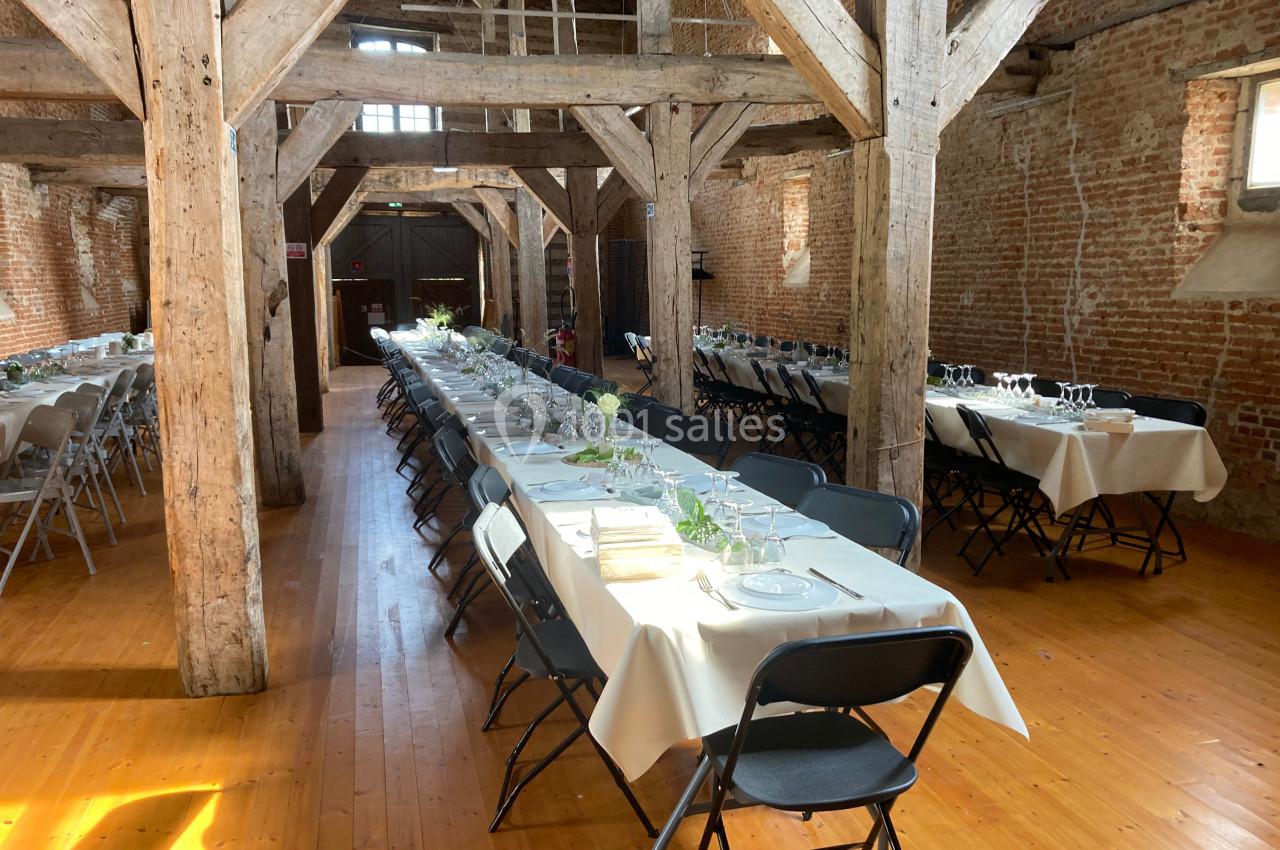Salle en bois avec tables rectangulaires dressées pour un repas, chaises noires et lumière naturelle entrant par les…