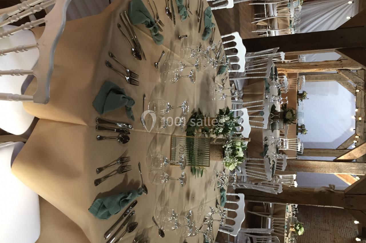 Table ronde dressée avec nappes beige, serviettes vertes, couverts, verres et centre de table floral dans une salle en bois.