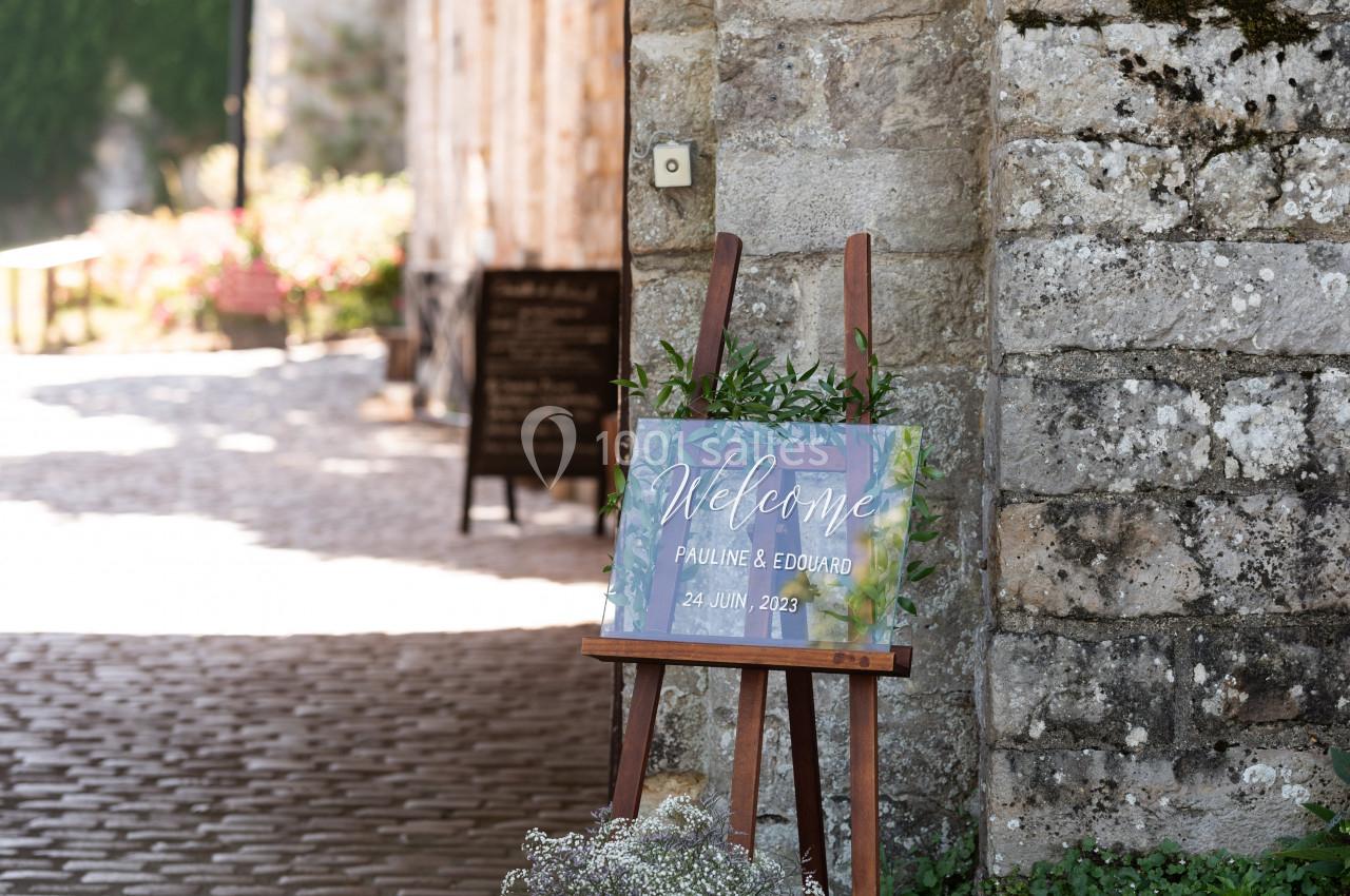 Panneau de bienvenue sur un chevalet en bois, entouré de verdure, devant un mur en pierre dans une cour pavée.