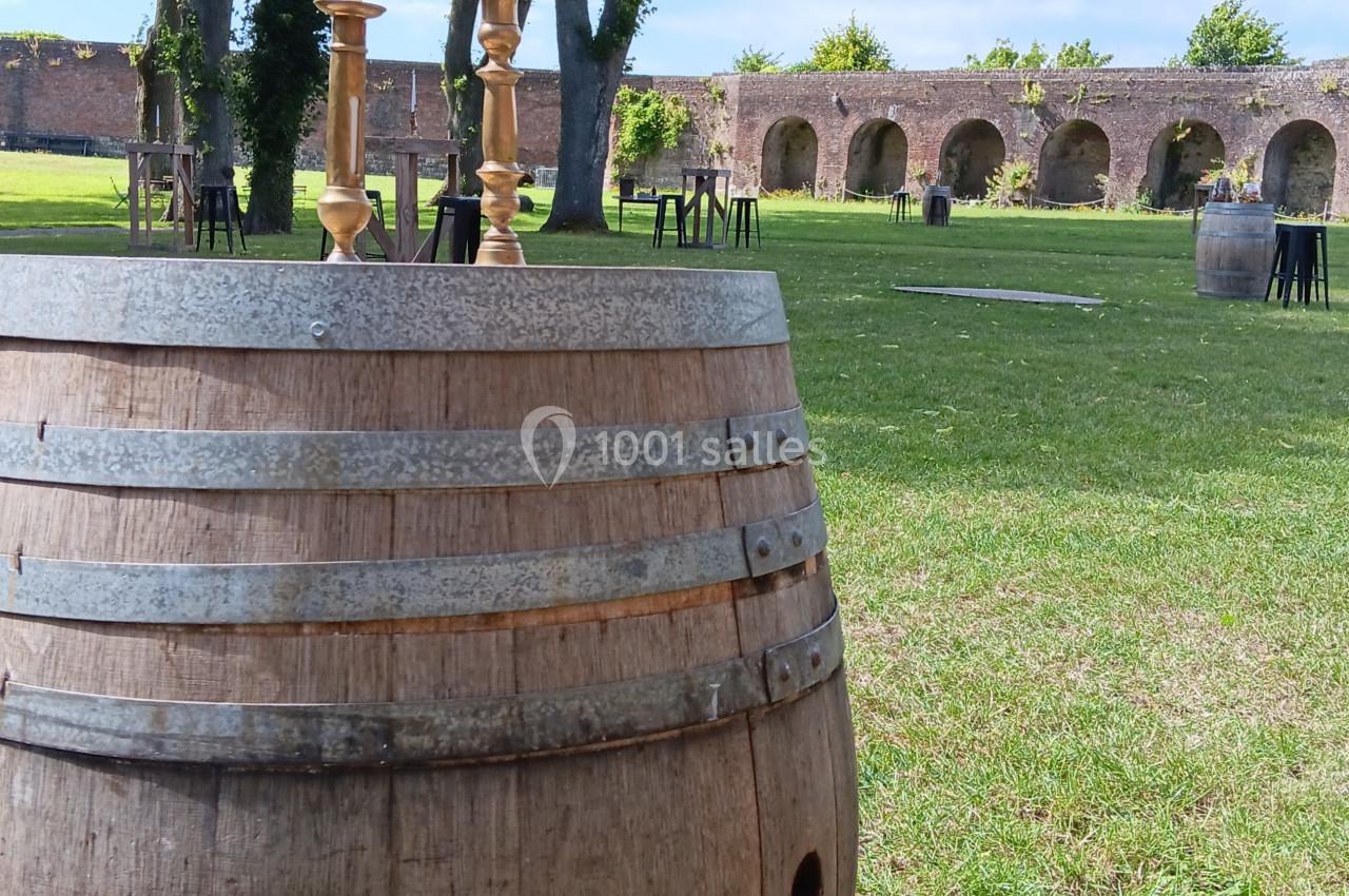 Tonneau en bois utilisé comme table avec des chandeliers, placé dans un jardin verdoyant devant des arches en pierre.
