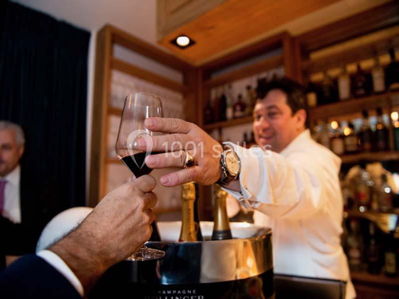 Un serveur tend un verre de vin rouge à un client devant un bar avec des bouteilles en arrière-plan.