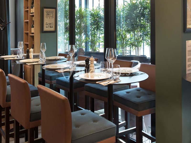 Salle de restaurant lumineuse avec tables dressées, chaises hautes et vue sur des plantes à travers de grandes fenêtres.