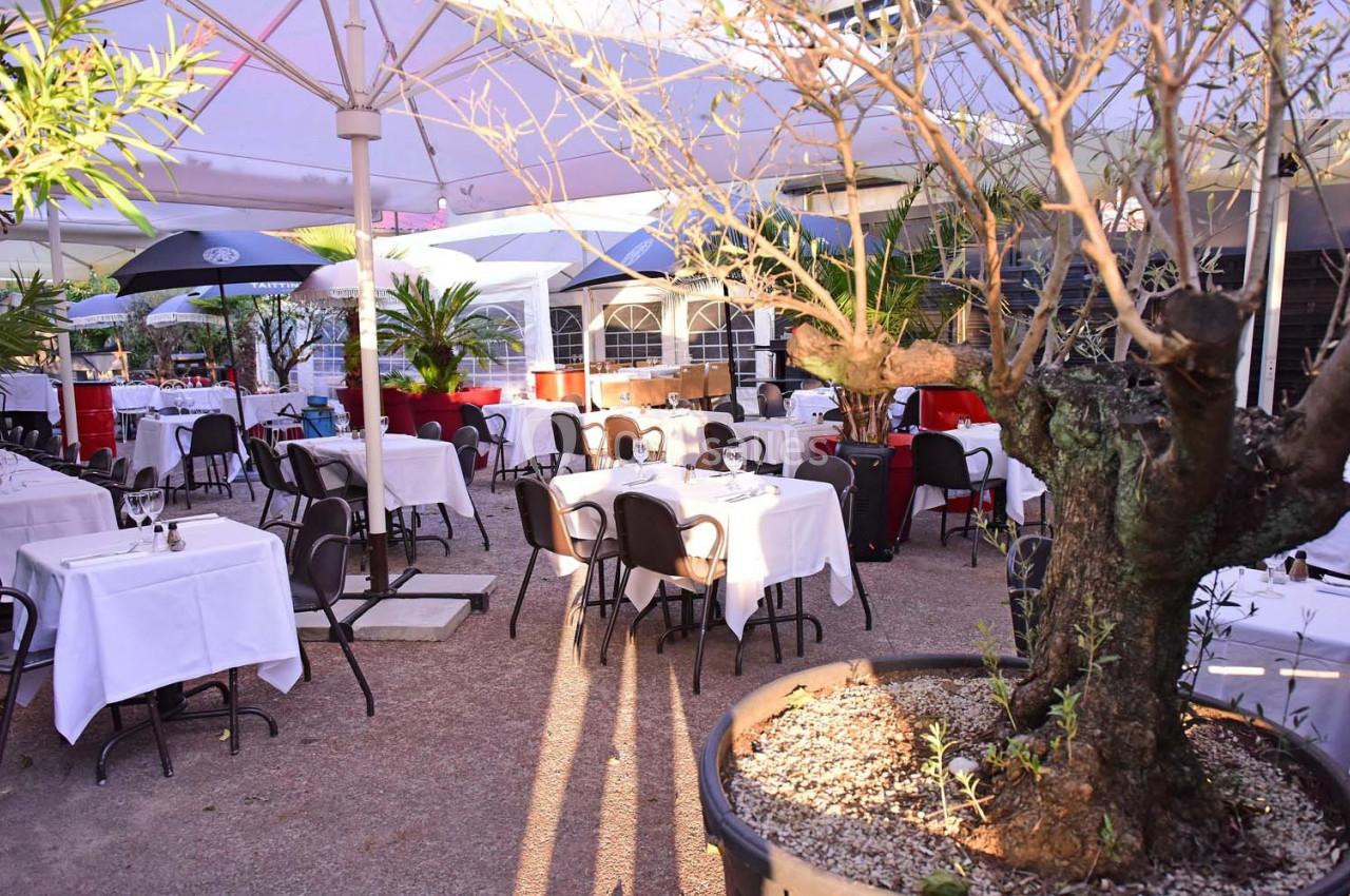 Terrasse d'un restaurant avec des tables dressées sous des parasols, entourée de plantes et d'arbres en pot.