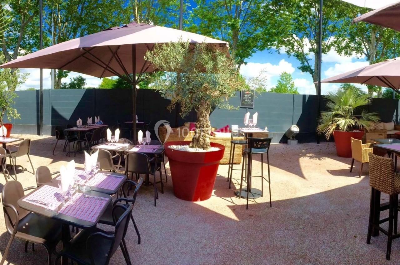 Terrasse extérieure avec tables dressées, chaises noires, parasols et plantes en pots sous un ciel dégagé.