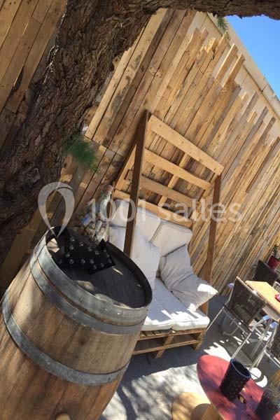 Banc en palettes avec coussins sous un arbre, près d'une barrique en bois et d'une table extérieure.