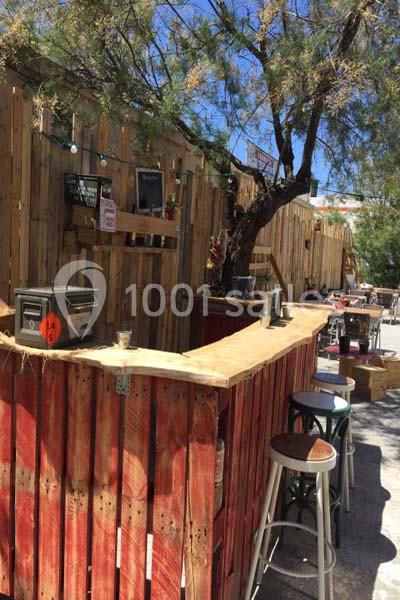 Bar extérieur en bois avec tabourets hauts, décoré de guirlandes lumineuses, situé sous un arbre par temps ensoleillé.