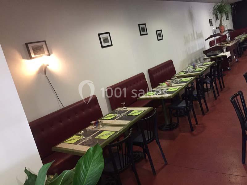 Salle de restaurant avec tables alignées, nappes vertes, chaises noires et banquettes rouges contre un mur décoré de cadres.