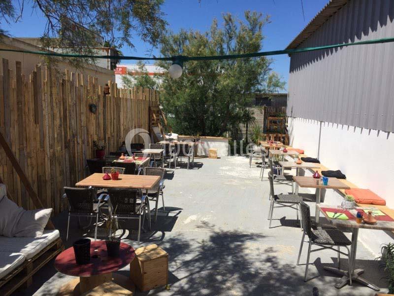 Terrasse extérieure avec tables en bois, chaises et coussins, entourée de palissades et végétation sous un ciel dégagé.