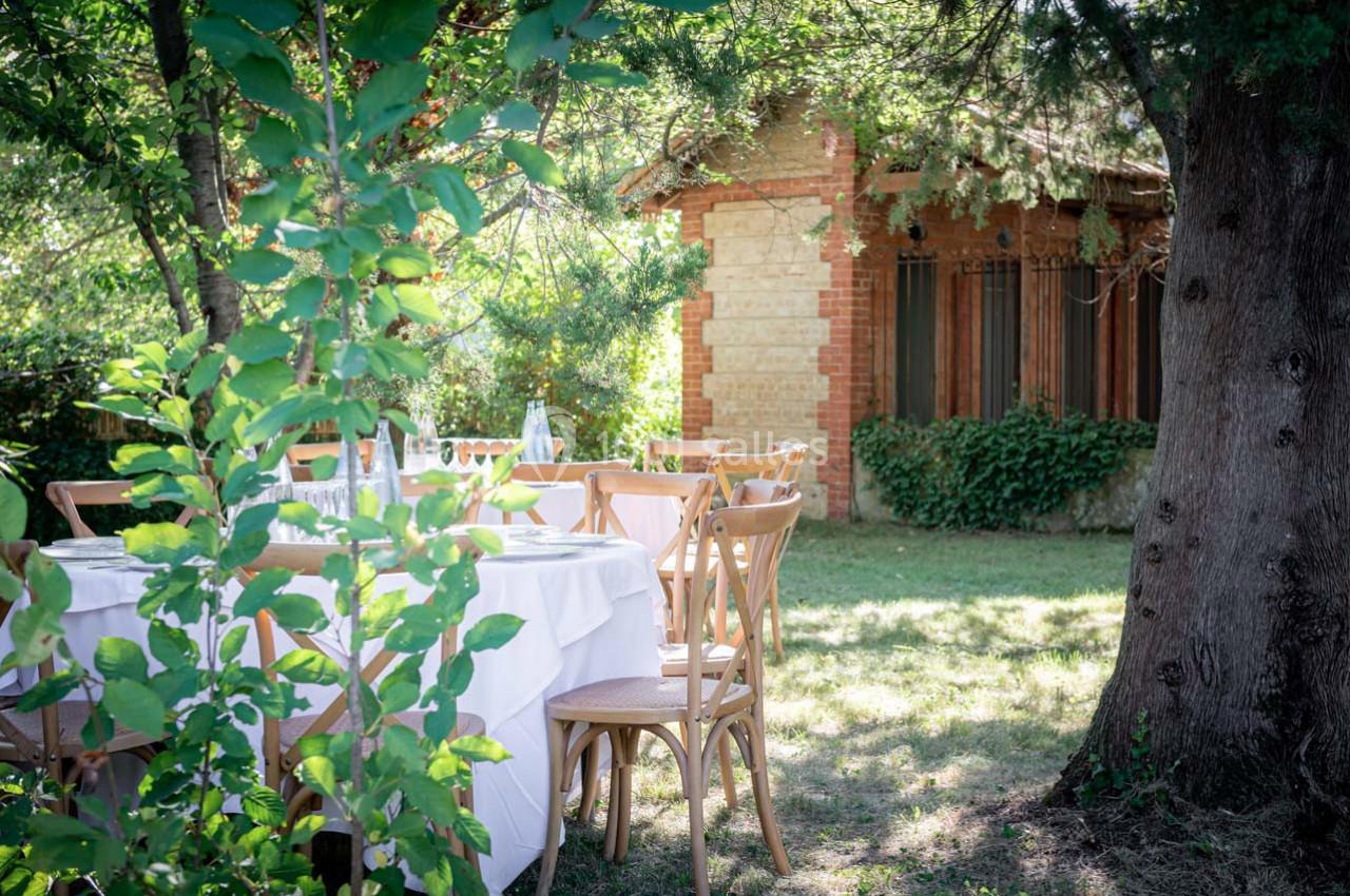Tables dressées avec nappes blanches et chaises en bois dans un jardin ombragé, près d'une maison en briques.