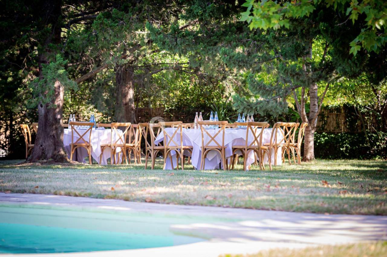 Tables dressées avec nappes blanches et chaises en bois disposées sous des arbres près d'une piscine dans un jardin.