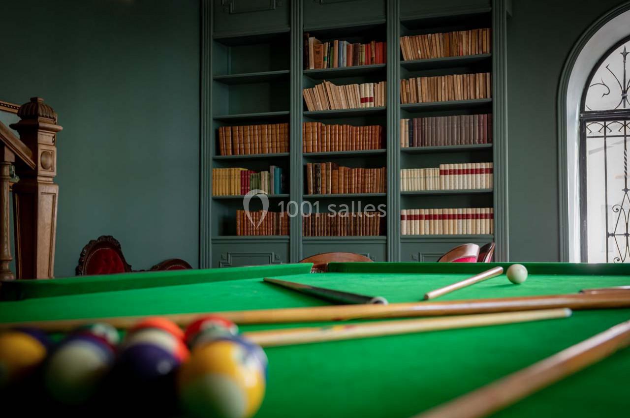 Table de billard avec des queues et des boules colorées au premier plan, bibliothèque en arrière-plan.