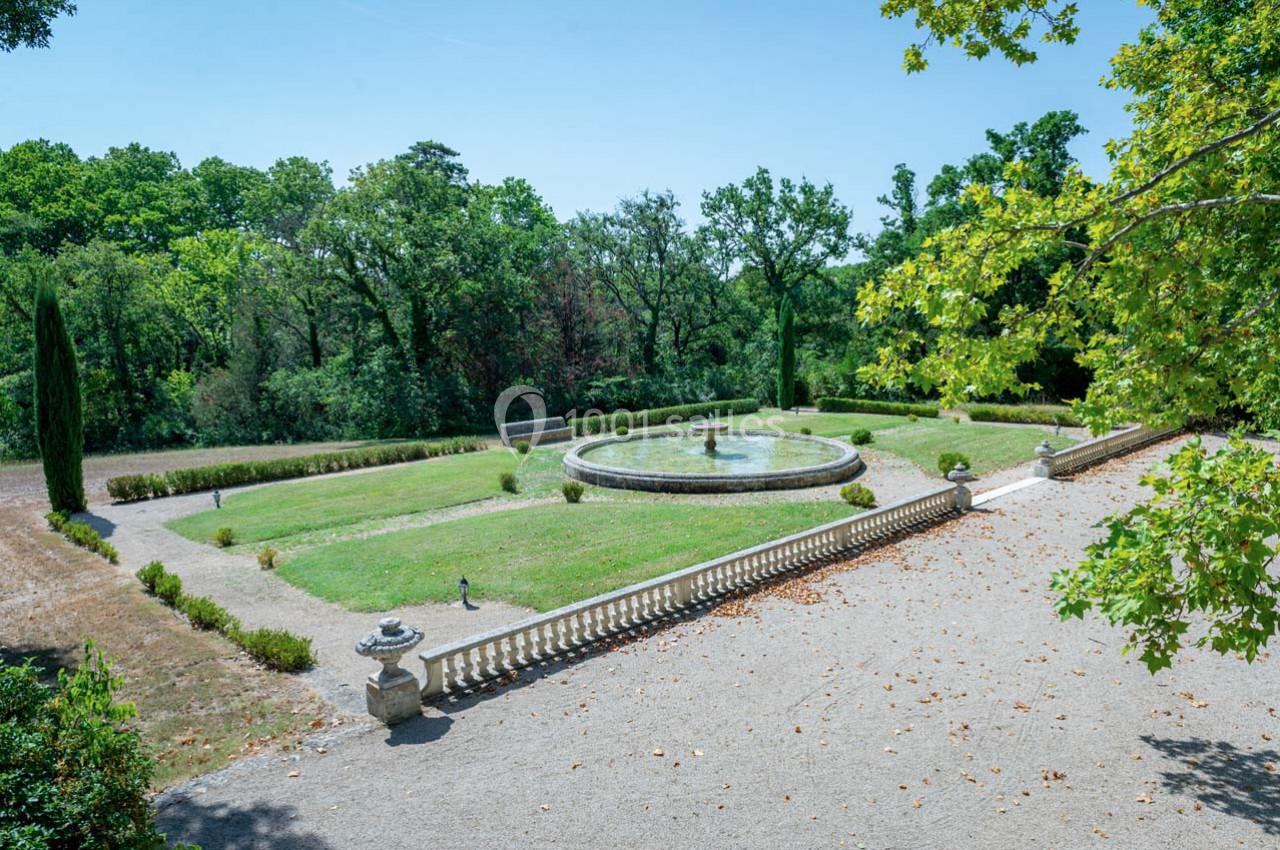 Jardin avec pelouse, fontaine centrale entourée de haies, balustrade en pierre et arbres en arrière-plan.