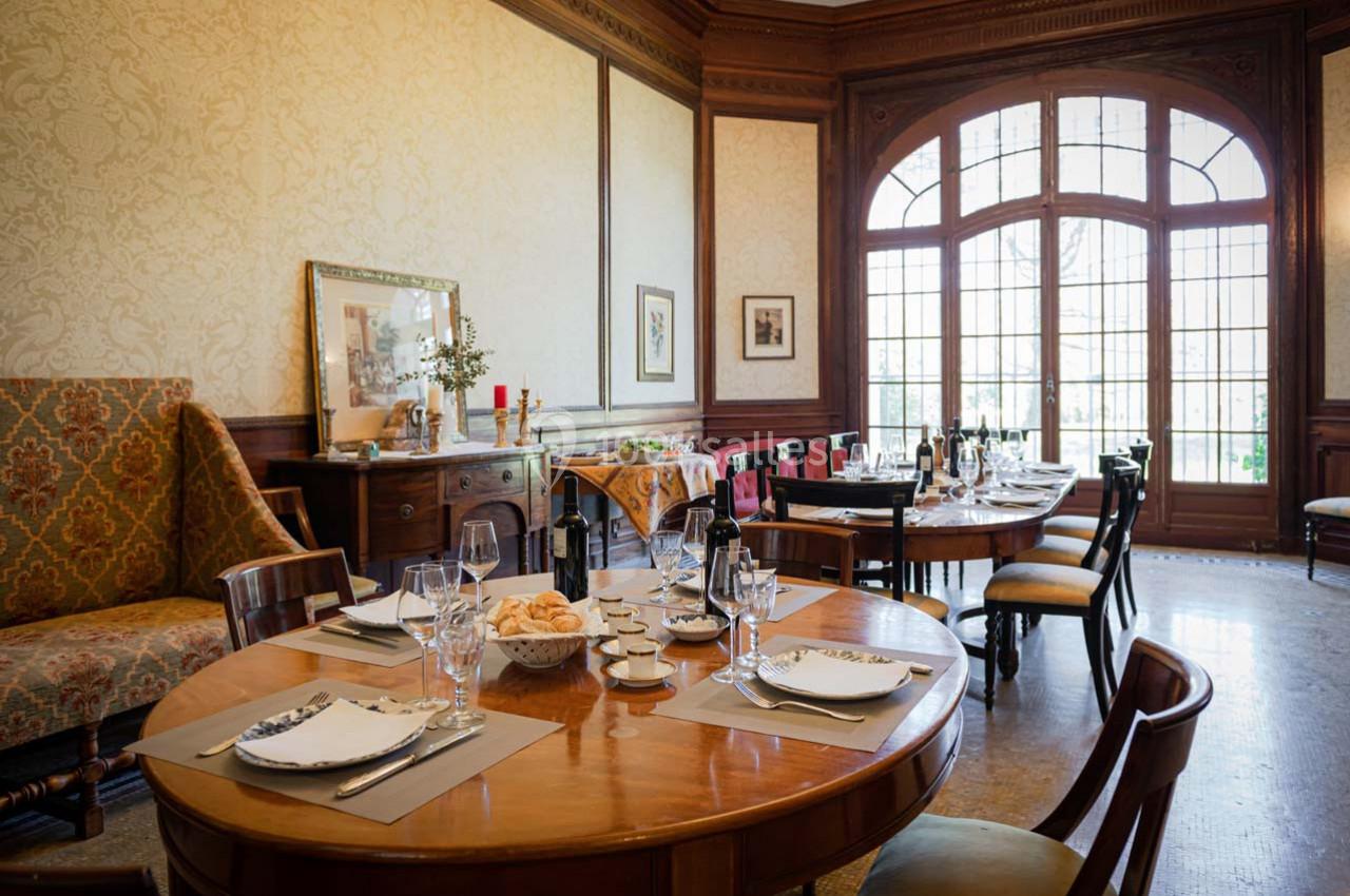 Salle à manger élégante avec tables dressées, vaisselle raffinée, grandes fenêtres et décoration classique en bois.