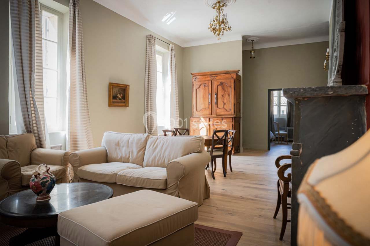 Salon lumineux avec canapé beige, table basse ronde, armoire en bois, chaises et grandes fenêtres avec rideaux clairs.