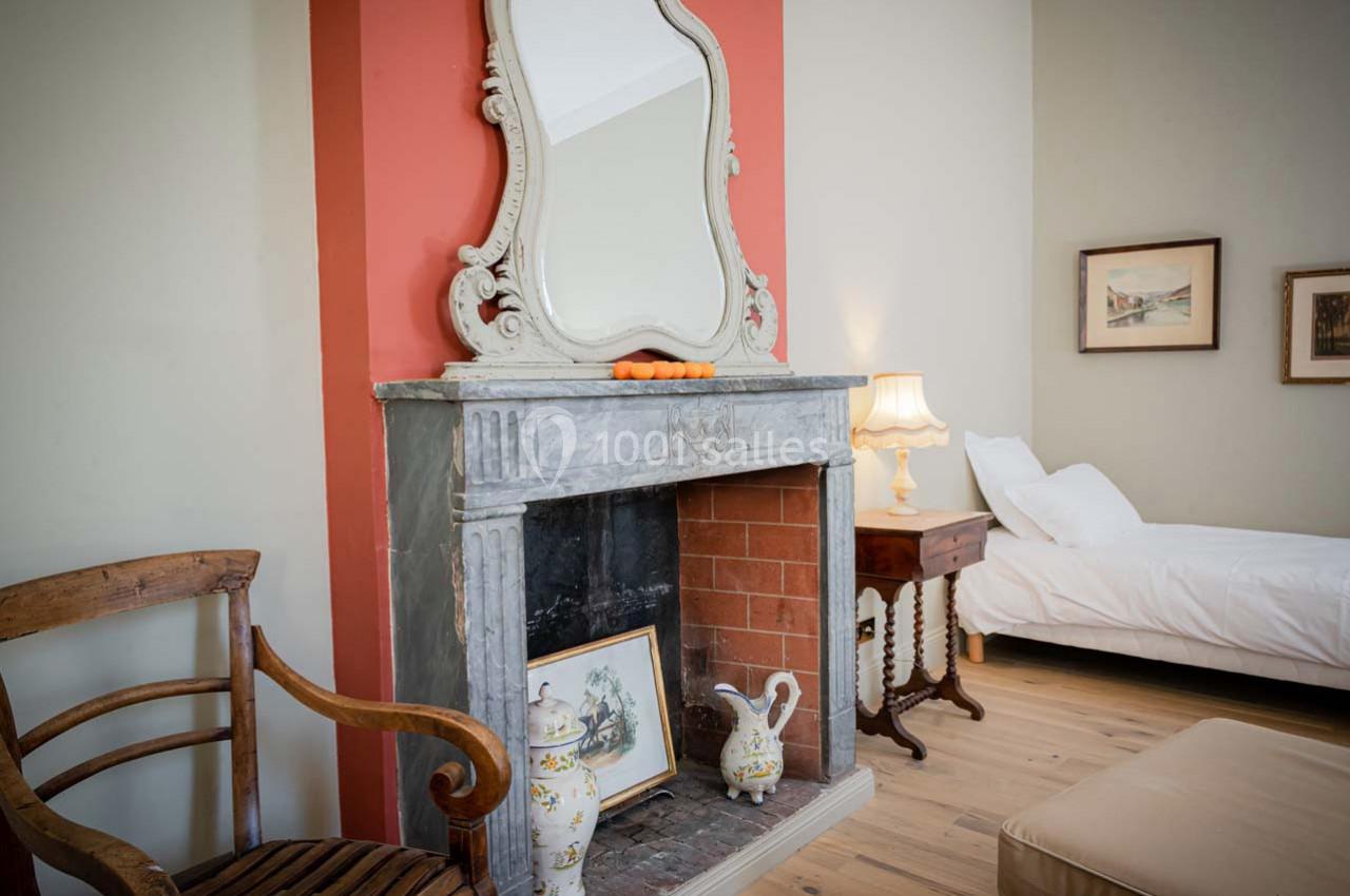 Cheminée en pierre avec miroir, objets décoratifs et chaise en bois dans une chambre lumineuse avec lit et lampe.