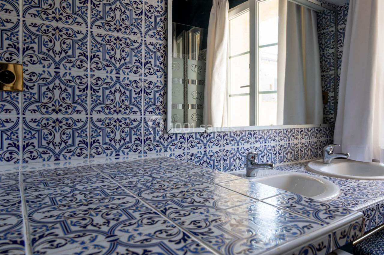 Salle de bain avec carrelage bleu et blanc à motifs, deux lavabos, un miroir et une fenêtre avec rideaux blancs.