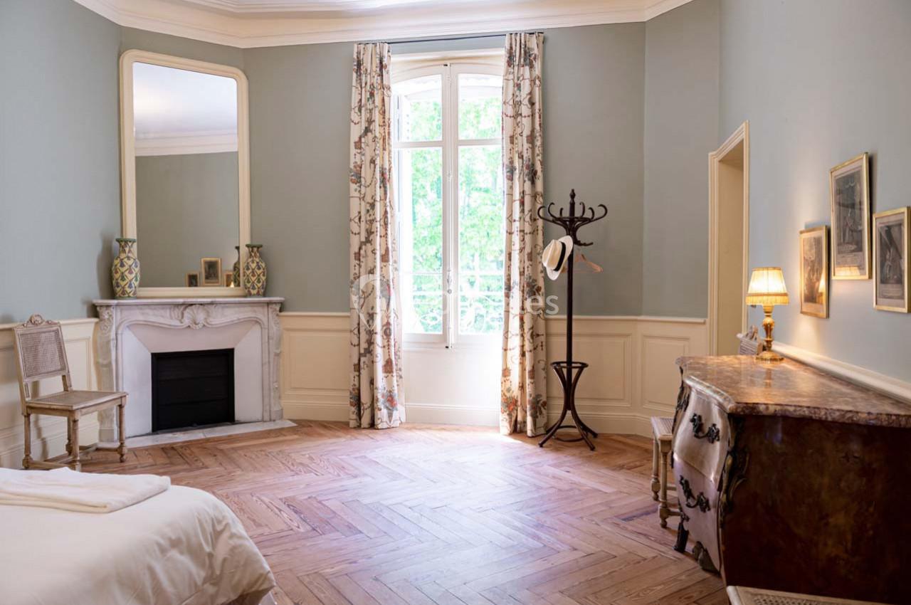 Chambre lumineuse avec parquet, cheminée décorative, fenêtre avec rideaux fleuris, et mobilier classique.