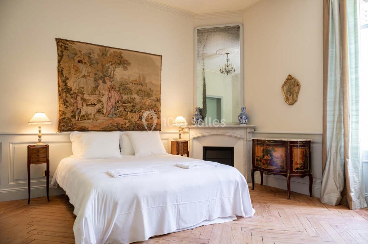 Chambre lumineuse avec lit double, tapisseries murales, cheminée décorative et mobilier ancien sur parquet en bois.