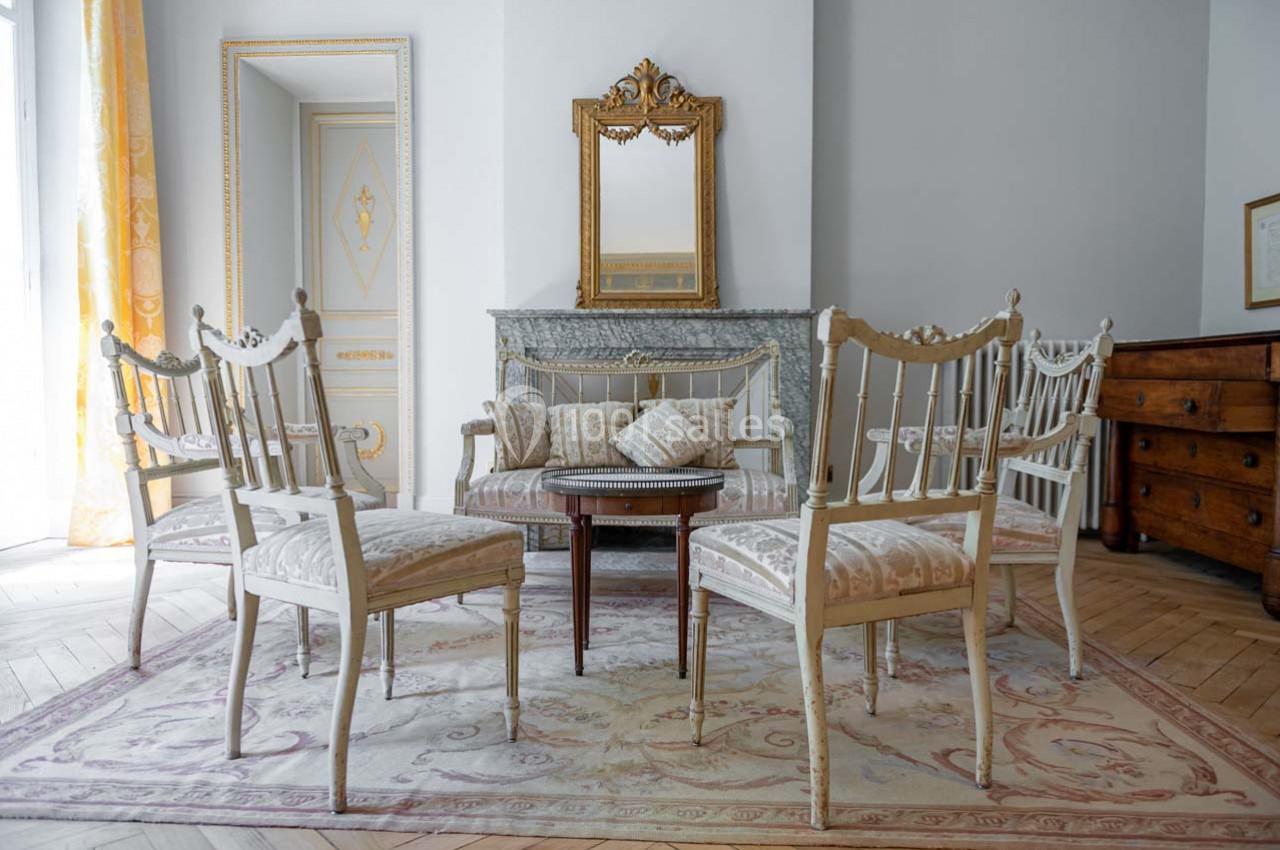 Mobilier ancien avec chaises et table en bois clair, disposés autour d'un tapis, devant une cheminée et un miroir doré.